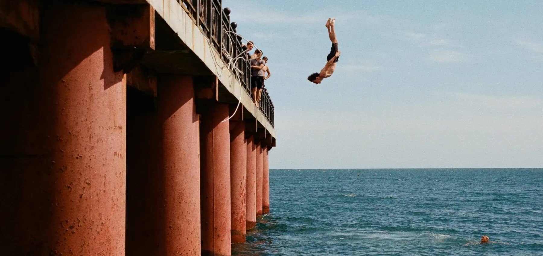 Next generation leadership, a young man somersaulting from a rusted iron pier into the sea while friends watch from the railing.