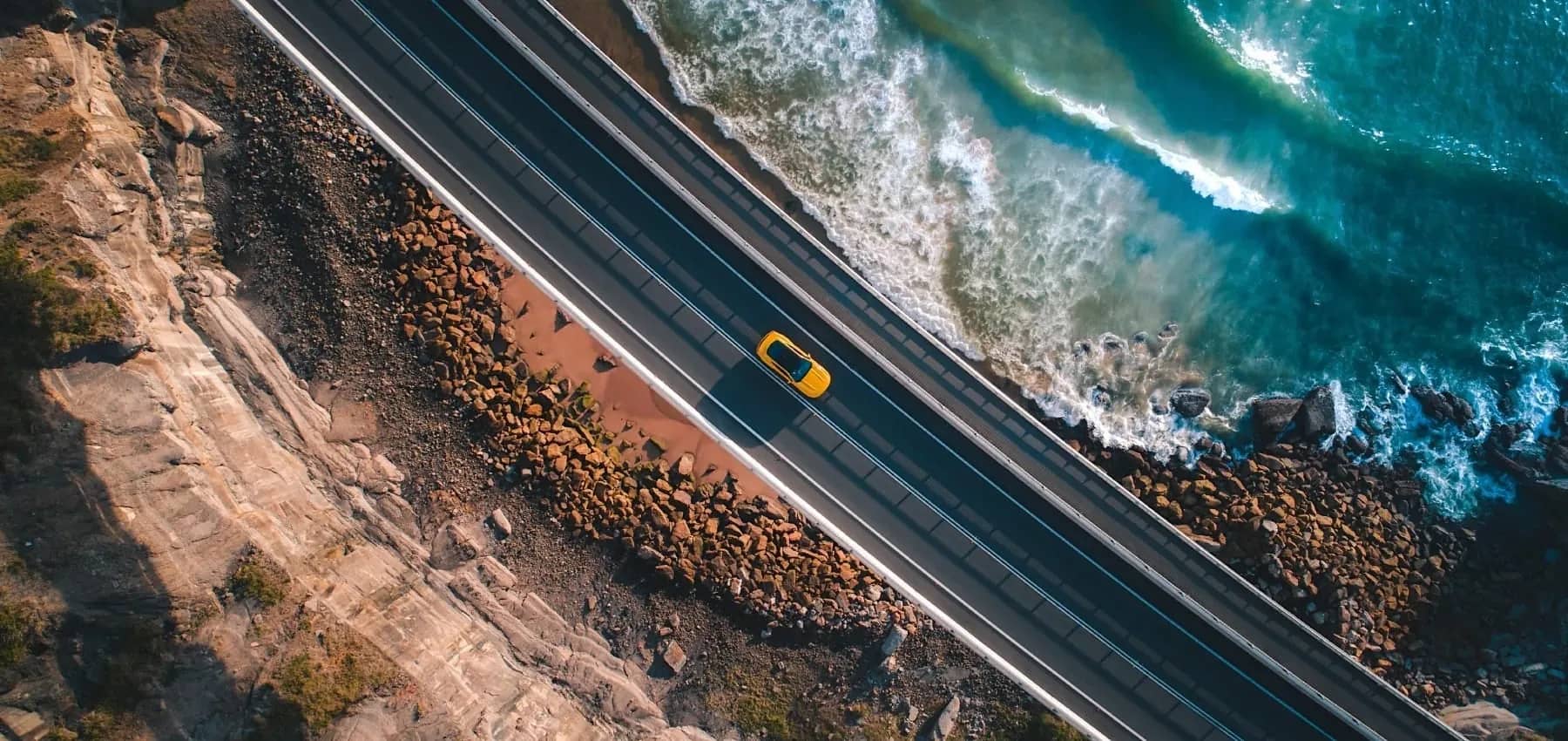 Long-term investing, an aerial view of a yellow car on a coastal road above the sea.