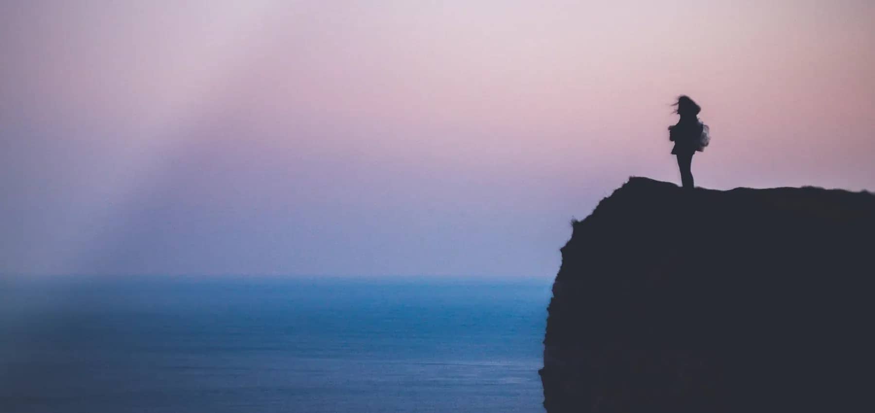Women on boards, a lone figure silhouetted on a clifftop above the sea at dusk.