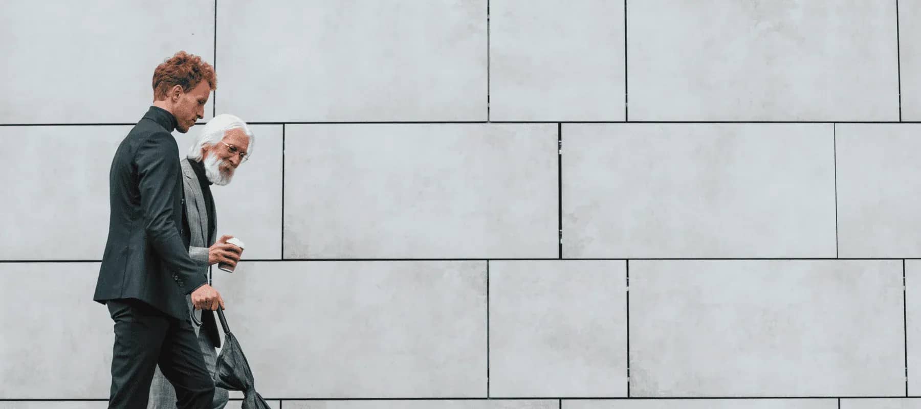 Next-gen principals, two men walking together past a long concrete wall.