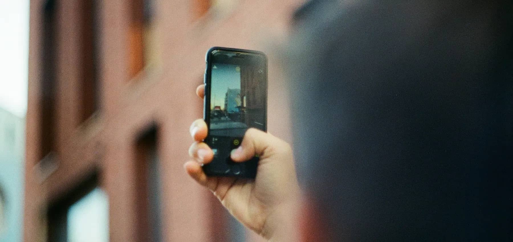 Next generation engagement, a hand holding up a smartphone to record, with an out-of-focus building behind.