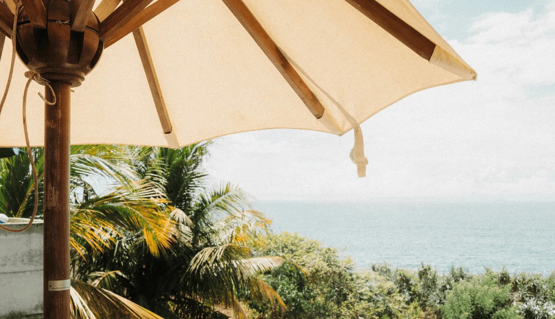 Lifestyle management concierge, a canvas parasol angled over palm fronds beside a pale sea.