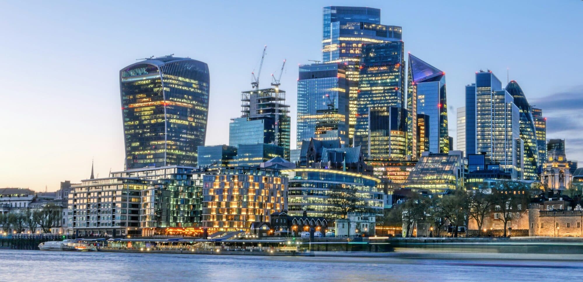 London financial district skyline at dusk with illuminated skyscrapers and River Thames in the foreground